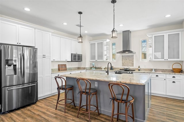 kitchen featuring wall chimney exhaust hood, hanging light fixtures, appliances with stainless steel finishes, a kitchen island with sink, and white cabinets