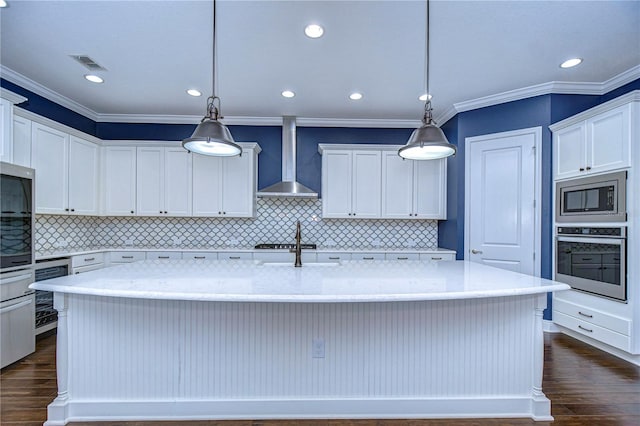 kitchen with appliances with stainless steel finishes, decorative light fixtures, a center island with sink, and wall chimney range hood