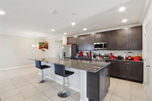 kitchen featuring appliances with stainless steel finishes, a kitchen island with sink, dark stone countertops, dark brown cabinets, and ornamental molding