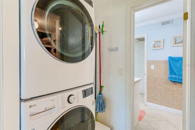 clothes washing area featuring light tile patterned flooring, stacked washer and clothes dryer, tile walls, a textured ceiling, and ornamental molding