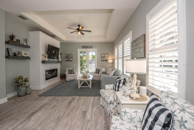 living room featuring a large fireplace, ceiling fan, a tray ceiling, light wood-type flooring, and french doors