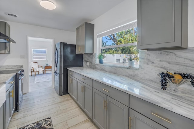 kitchen with gray cabinetry, stainless steel appliances, light stone countertops, light hardwood / wood-style floors, and backsplash