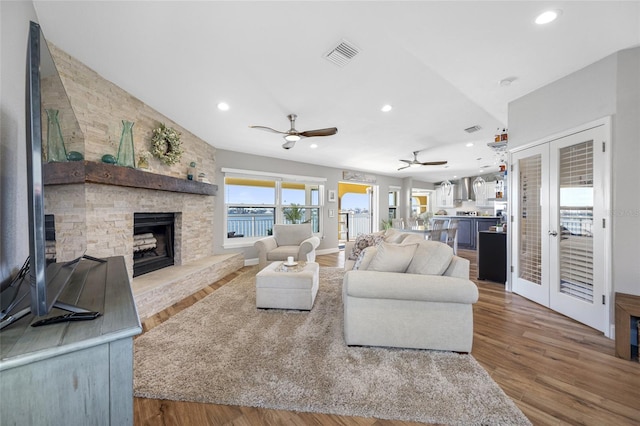 living room featuring french doors, a stone fireplace, a water view, hardwood / wood-style flooring, and ceiling fan