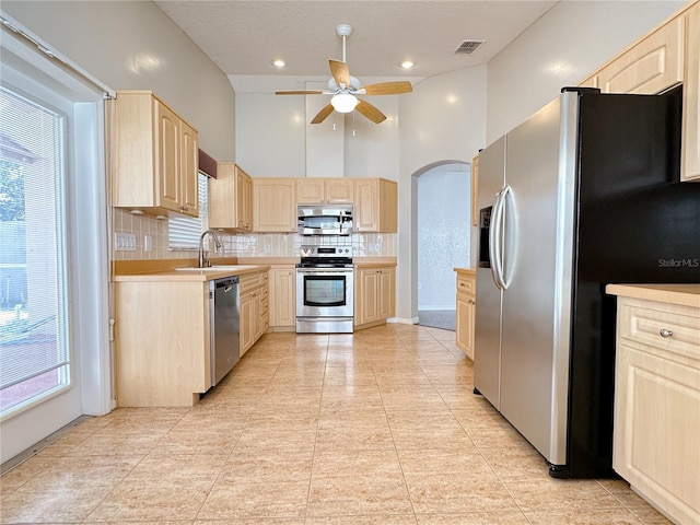 kitchen featuring stainless steel appliances, light brown cabinetry, decorative backsplash, and a high ceiling