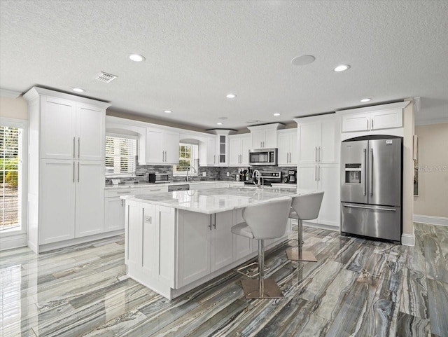 kitchen with appliances with stainless steel finishes, white cabinetry, plenty of natural light, light stone countertops, and an island with sink