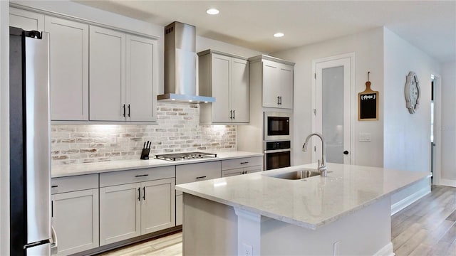 kitchen featuring appliances with stainless steel finishes, an island with sink, sink, light stone countertops, and wall chimney exhaust hood