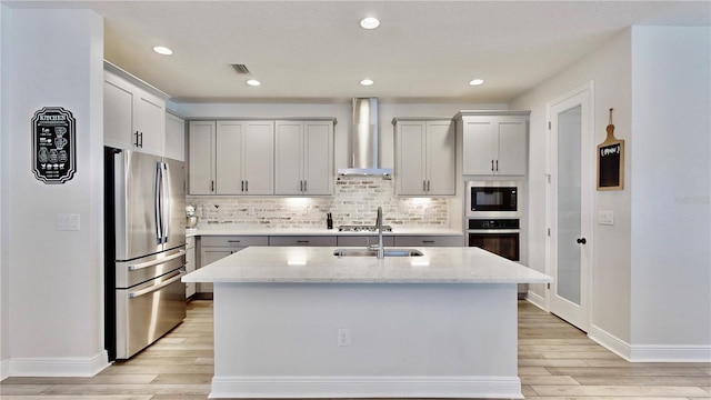 kitchen with stainless steel appliances, light stone countertops, a kitchen island with sink, and wall chimney range hood