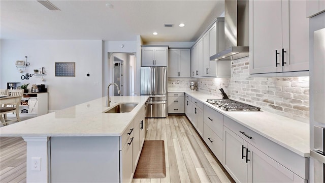 kitchen featuring wall chimney exhaust hood, sink, an island with sink, stainless steel appliances, and light stone countertops