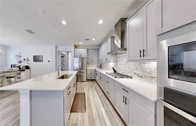 kitchen featuring sink, appliances with stainless steel finishes, a center island with sink, decorative backsplash, and wall chimney exhaust hood