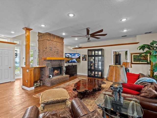 living room with a brick fireplace, light hardwood / wood-style flooring, a textured ceiling, ceiling fan, and decorative columns