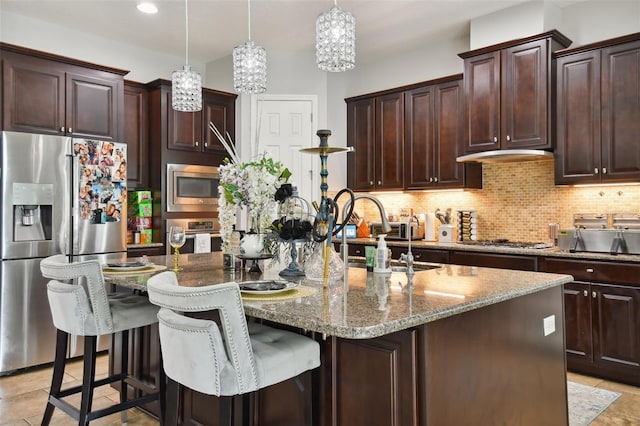 kitchen with dark stone countertops, stainless steel appliances, tasteful backsplash, an island with sink, and decorative light fixtures