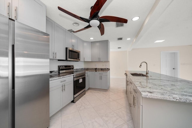 kitchen featuring sink, ceiling fan, stainless steel appliances, light stone counters, and an island with sink