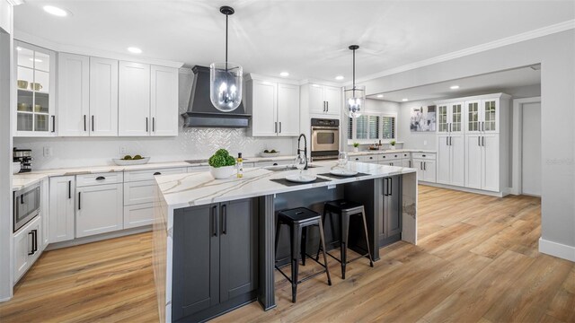 kitchen with pendant lighting, wall chimney range hood, appliances with stainless steel finishes, an island with sink, and white cabinets