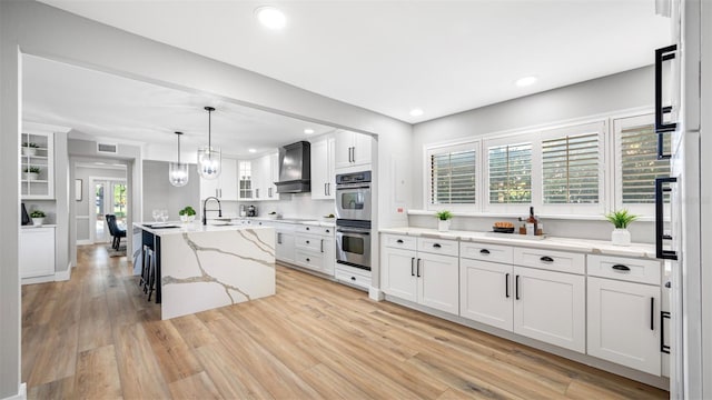 kitchen featuring pendant lighting, double oven, white cabinets, a kitchen island with sink, and custom range hood