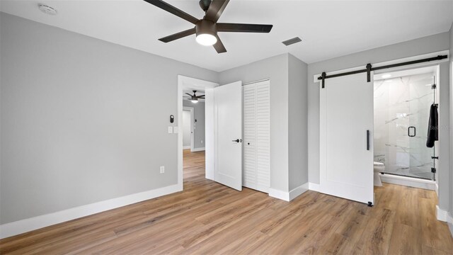 unfurnished bedroom with ceiling fan, ensuite bathroom, light hardwood / wood-style floors, a barn door, and a closet