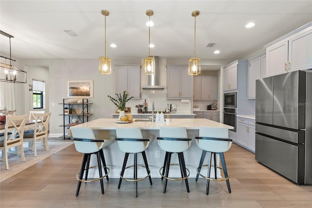 kitchen featuring appliances with stainless steel finishes, a breakfast bar, decorative light fixtures, a kitchen island with sink, and wall chimney range hood