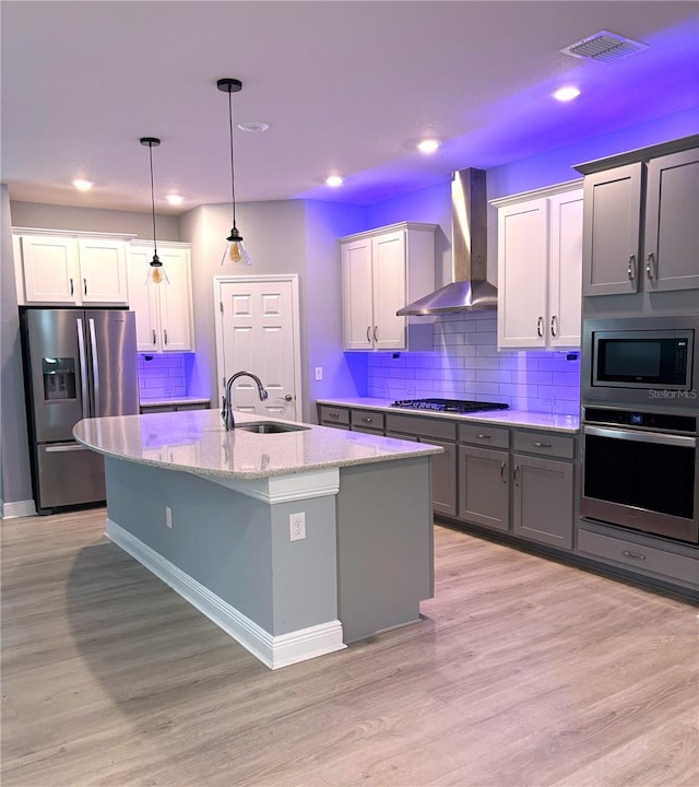 kitchen featuring appliances with stainless steel finishes, sink, wall chimney range hood, hanging light fixtures, and a center island with sink