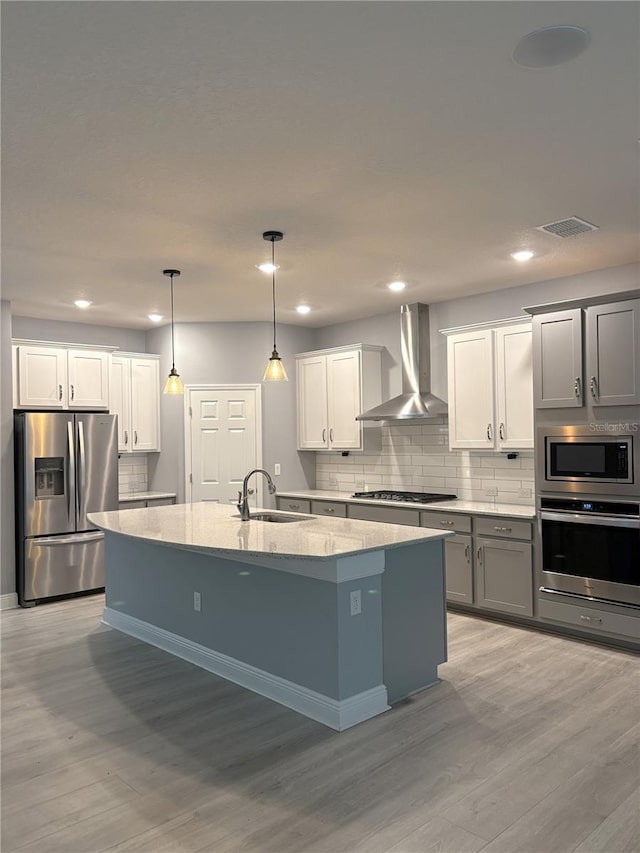 kitchen featuring a center island with sink, appliances with stainless steel finishes, pendant lighting, light stone countertops, and wall chimney range hood