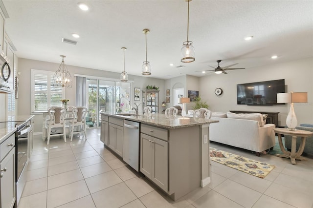 kitchen with gray cabinets, appliances with stainless steel finishes, pendant lighting, an island with sink, and light stone countertops