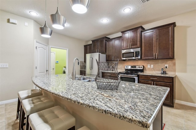 kitchen with a kitchen island with sink, backsplash, stainless steel appliances, light stone counters, and decorative light fixtures