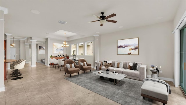 tiled living room featuring ceiling fan with notable chandelier, a tray ceiling, and decorative columns