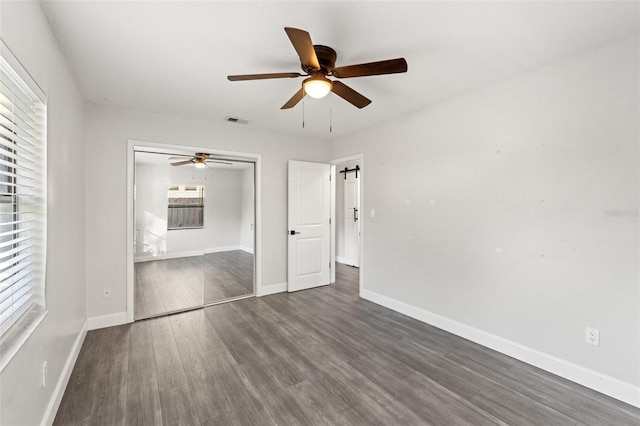 unfurnished bedroom featuring ceiling fan, a barn door, dark hardwood / wood-style flooring, and a closet