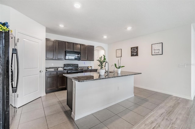 kitchen with dark stone countertops, a center island with sink, arched walkways, black appliances, and dark brown cabinets