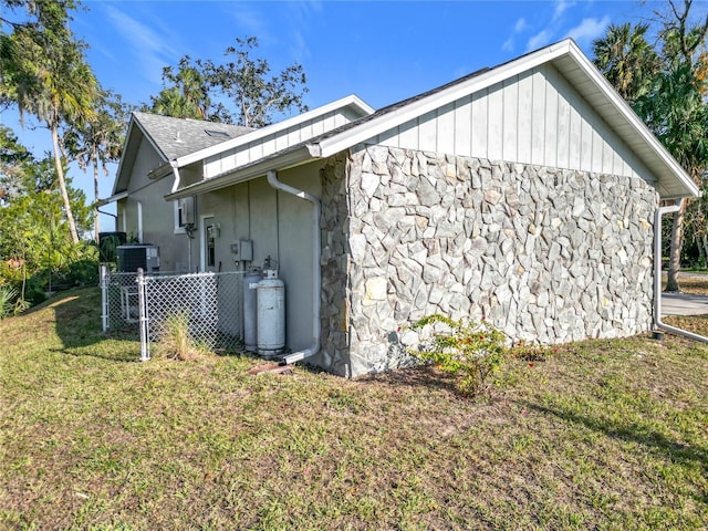 view of side of property with central AC, fence, stone siding, a lawn, and roof with shingles