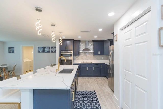 kitchen with pendant lighting, sink, a center island with sink, separate washer and dryer, and wall chimney exhaust hood