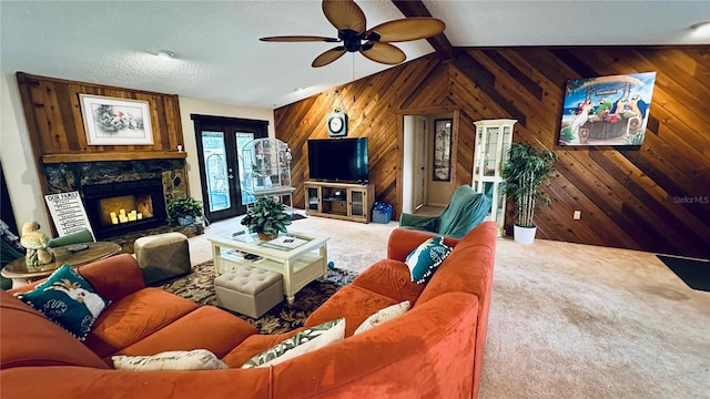 carpeted living room with ceiling fan, wooden walls, a stone fireplace, vaulted ceiling, and french doors