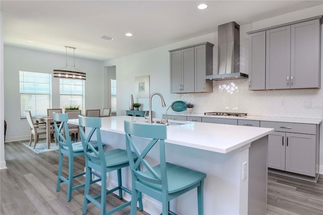 kitchen featuring wall chimney exhaust hood, sink, a center island with sink, gray cabinets, and decorative backsplash