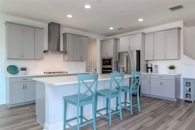 kitchen featuring appliances with stainless steel finishes, a breakfast bar area, light hardwood / wood-style floors, a center island with sink, and wall chimney exhaust hood