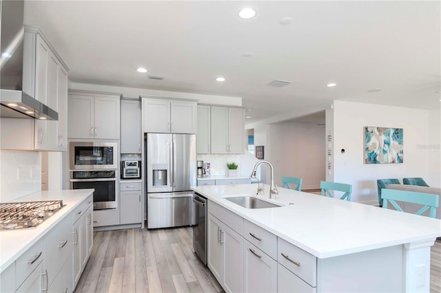 kitchen featuring wall chimney range hood, sink, light hardwood / wood-style flooring, appliances with stainless steel finishes, and a kitchen island with sink