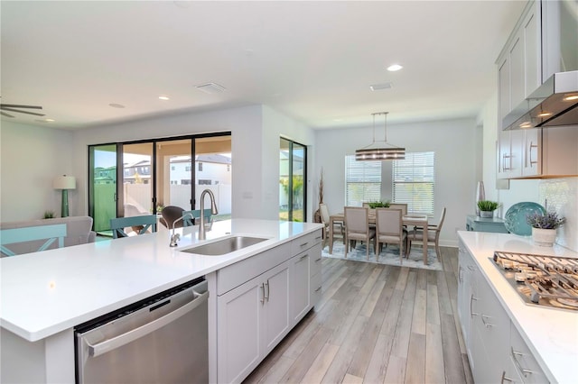 kitchen with wall chimney range hood, sink, a kitchen island with sink, stainless steel appliances, and decorative light fixtures