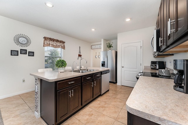 kitchen with sink, light tile patterned floors, appliances with stainless steel finishes, dark brown cabinetry, and an island with sink