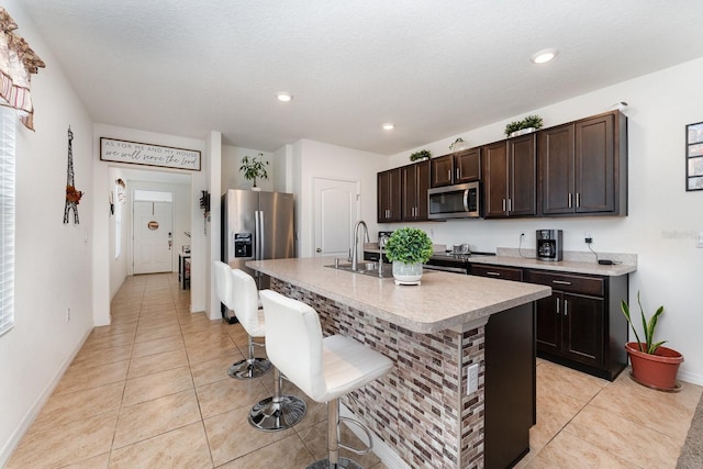 kitchen featuring a breakfast bar, a kitchen island with sink, stainless steel appliances, dark brown cabinetry, and light tile patterned flooring