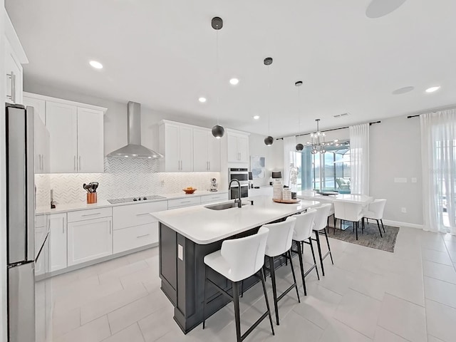 kitchen with hanging light fixtures, an island with sink, white cabinets, and wall chimney exhaust hood