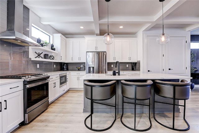 kitchen featuring wall chimney exhaust hood, appliances with stainless steel finishes, a center island with sink, and white cabinets