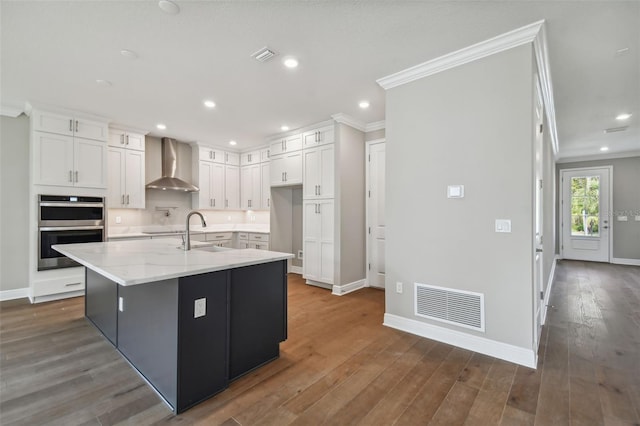 kitchen featuring white cabinetry, a kitchen island with sink, wall chimney range hood, and stainless steel double oven