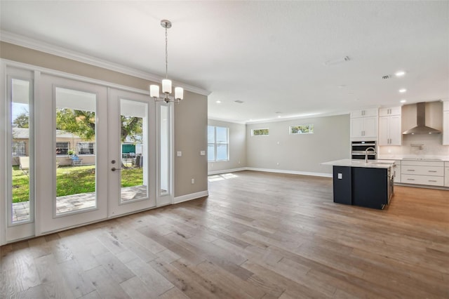 kitchen featuring an island with sink, white cabinets, hanging light fixtures, wall chimney range hood, and light wood-type flooring