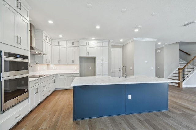 kitchen with white cabinetry, a kitchen island with sink, wall chimney range hood, and double oven