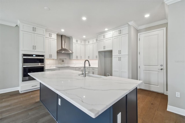 kitchen with a center island with sink, white cabinetry, wall chimney range hood, and double oven