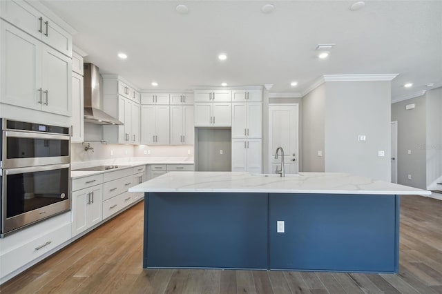 kitchen featuring double oven, white cabinets, wall chimney exhaust hood, and a large island with sink