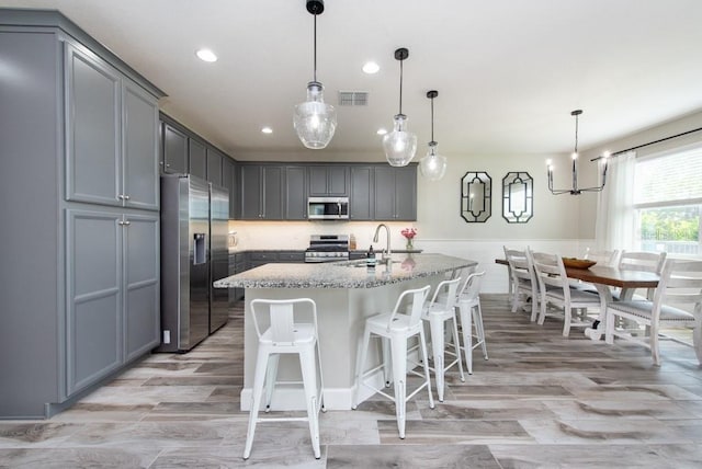 kitchen featuring appliances with stainless steel finishes, sink, hanging light fixtures, light stone counters, and a center island with sink