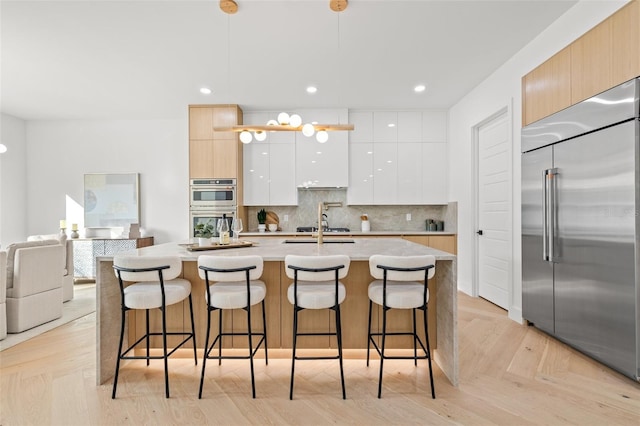 kitchen with a center island with sink, light brown cabinets, hanging light fixtures, stainless steel appliances, and white cabinets