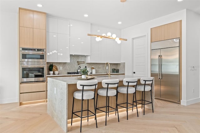 kitchen with white cabinetry, decorative light fixtures, appliances with stainless steel finishes, an island with sink, and light parquet flooring