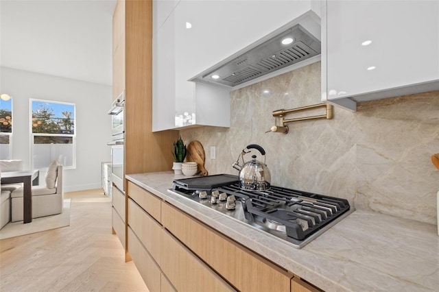 kitchen with light brown cabinetry, exhaust hood, tasteful backsplash, stainless steel gas stovetop, and light parquet floors