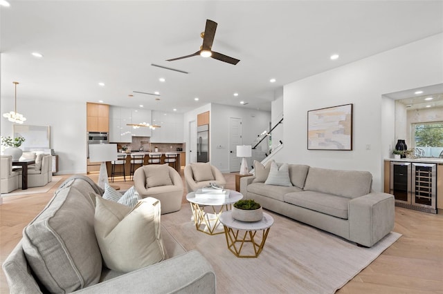 living room with wine cooler, ceiling fan with notable chandelier, and light wood-type flooring