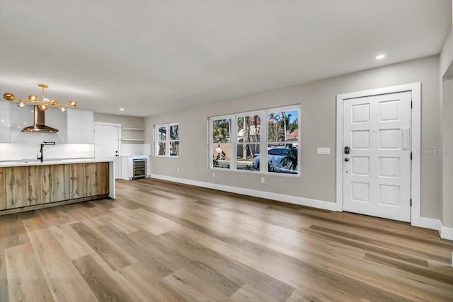 unfurnished living room featuring an inviting chandelier, beverage cooler, sink, and light hardwood / wood-style flooring