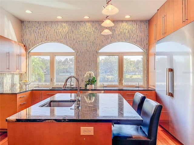 kitchen featuring a kitchen island with sink, hanging light fixtures, dark stone countertops, a wealth of natural light, and stainless steel built in fridge